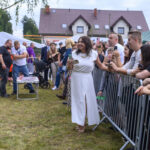 Na zdjęciu jest kobieta w białej sukni stojąca na trawie. Obok niej są ludzie za metalowymi barierkami. W tle widać dom i drzewo.