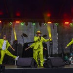 Trzech mężczyzn w neonowych, zielonych strojach występuje na scenie. Tło jest ciemne, oświetlone kolorowymi światłami. Scena ustawiona jest pod zadaszeniem.