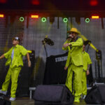 Na scenie zespół w jaskrawych zielonych strojach. Śpiewający artysta w żółtym kapeluszu. Tło z kolorowymi światłami.