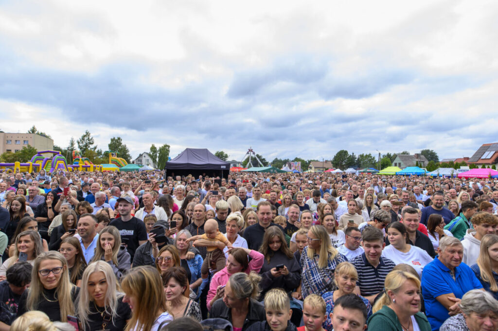 Tłum ludzi na zewnątrz podczas festiwalu. Tła w kolorze nieba z chmurami, kolorowe namioty i zjeżdżalnie. Uczestnicy uśmiechają się, atmosfera wesoła.