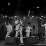 Czterech mężczyzn w białych garniturach występuje na ciemnej scenie. Jeden z nich śpiewa do mikrofonu. Scena oświetlona reflektorami.