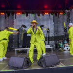 Na scenie są cztery osoby w jasnozielonych garniturach. Tło to czarny materiał z widocznymi światłami. Na scenie widoczne są instrumenty muzyczne.