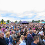 Na zdjęciu widoczny jest tłum ludzi na zewnątrz, na tle białego, pochmurnego nieba. Ludzie w różnym wieku stoją blisko siebie, a z tyłu widać kolorowe stragany. Na horyzoncie widoczne są drzewa i budynki