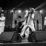 Trzech artystów w białych garniturach występuje na scenie. Jeden z nich śpiewa do mikrofonu. Tło stanowi oświetlony reflektorami ekran.