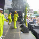 Na scenie trzech artystów w jasnozielonych strojach. Tło to duża widownia na świeżym powietrzu. W tle drzewa i namioty.