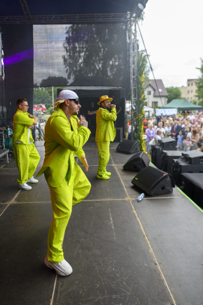 Trzech mężczyzn w neonowych zielonych strojach występuje na scenie. Tłum ludzi ogląda ich koncert na świeżym powietrzu. Tło jest ciemne, a w oddali widać drzewa i budynki.