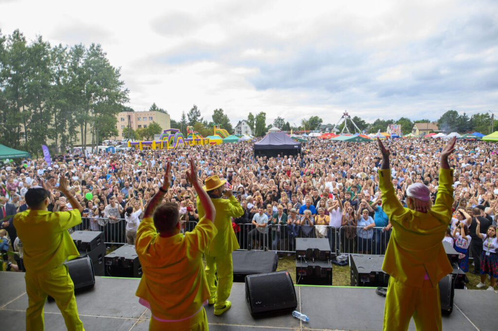 Koncert na świeżym powietrzu, czterech artystów w żółtych strojach, tłum ludzi, kolorowe namioty i drzewa w tle.