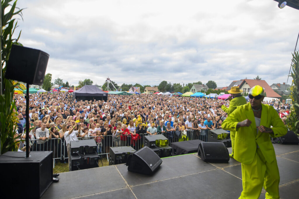 Koncert w plenerze z tłumem ludzi i sceną na pierwszym planie. Mężczyzna w jaskrawożółtym stroju na scenie. Chmury na niebie.