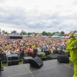Koncert w plenerze z tłumem ludzi i sceną na pierwszym planie. Mężczyzna w jaskrawożółtym stroju na scenie. Chmury na niebie.