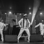 Trzech mężczyzn w jasnych strojach występuje na scenie w świetle reflektorów. Środkowy ma kapelusz. Tło ciemne, scena w czerni i bieli.
