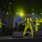 Na scenie trzech mężczyzn w zielonych garniturach, w tle sprzęt muzyczny. Scena oświetlona żółtymi i niebieskimi światłami.