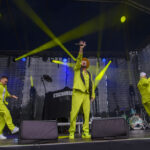 Na scenie trzech mężczyzn w neonowo-żółtych strojach. Tło jest ciemne z żółtymi światłami. Na środku mężczyzna trzyma mikrofon.