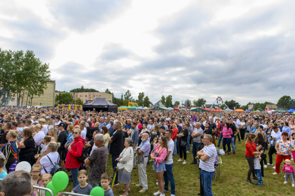 Duża grupa ludzi na trawniku podczas festiwalu na świeżym powietrzu. W tle kolorowe namioty i drzewa. Niebo jest pochmurne.