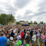 Tłum ludzi na zewnątrz, tło z drzewami i chmurami. Wiele osób trzyma zielone balony. Scena festynu z namiotami.