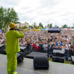 Mężczyzna w jaskrawym zielonym garniturze i żółtym kapeluszu śpiewa na scenie. Przed nim tłum widzów na zielonym tle trawy. W tle widać drzewa i namioty na festynie.