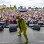 Na scenie stoi artysta w jaskrawożółtym garniturze. Tło to tłum ludzi na plenerowym wydarzeniu. Po lewej kolorowe atrakcje i namioty.