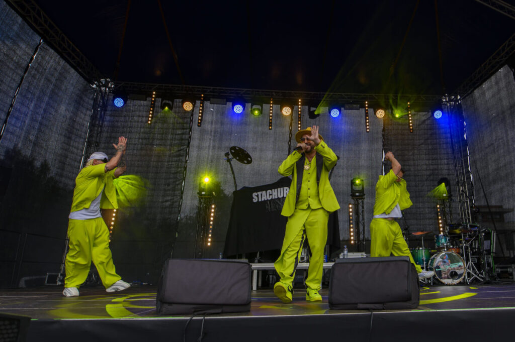 Na scenie trzech artystów w jaskrawych, limonkowych strojach. Tło ciemne, z kolorowymi światłami sceny. Instrumenty i sprzęt DJ-a w tle.