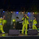 Na scenie trzech artystów w jaskrawych, limonkowych strojach. Tło ciemne, z kolorowymi światłami sceny. Instrumenty i sprzęt DJ-a w tle.