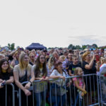 Grupa ludzi za barierkami na koncercie plenerowym. W tle jasne niebo i drzewa. Dominują jasne i naturalne kolory.