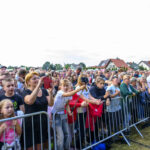 Tłum ludzi stoi za barierką na zewnątrz. Większość osób trzyma telefony, robiąc zdjęcia. W tle domy i drzewa pod jasnym niebem.