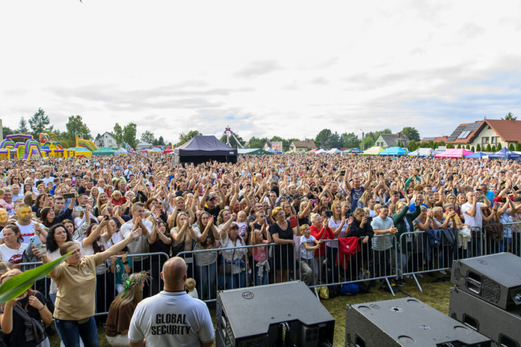 Na zdjęciu tłum ludzi na festiwalu pod gołym niebem. Tło to kolorowe namioty i drzewa. Przód sceny zabezpieczany przez ochroniarzy.