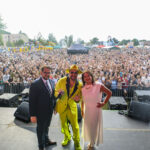 Na zdjęciu scena z trojgiem ludzi; mężczyzna w żółtym garniturze, kobieta w białej sukience, tłum ludzi w tle. Kolorowe dmuchańce po lewej.