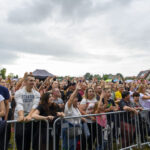 Na zdjęciu widać tłum ludzi na koncercie na świeżym powietrzu. Przeważają kolory jasne i naturalne, a w tle są domy i drzewa. Niebo jest pochmurne.