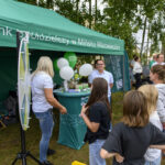 Ludzie stoją przed zielonym namiotem banku. Na stoliku balony i materiały promocyjne. Tło: drzewa i tłum.