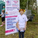 Osoba w białym kitlu i wianku kwiatów stoi przed plakatem usług medycznych. Tło z drzewami i trawa. Plakat zawiera różnokolorowe napisy.