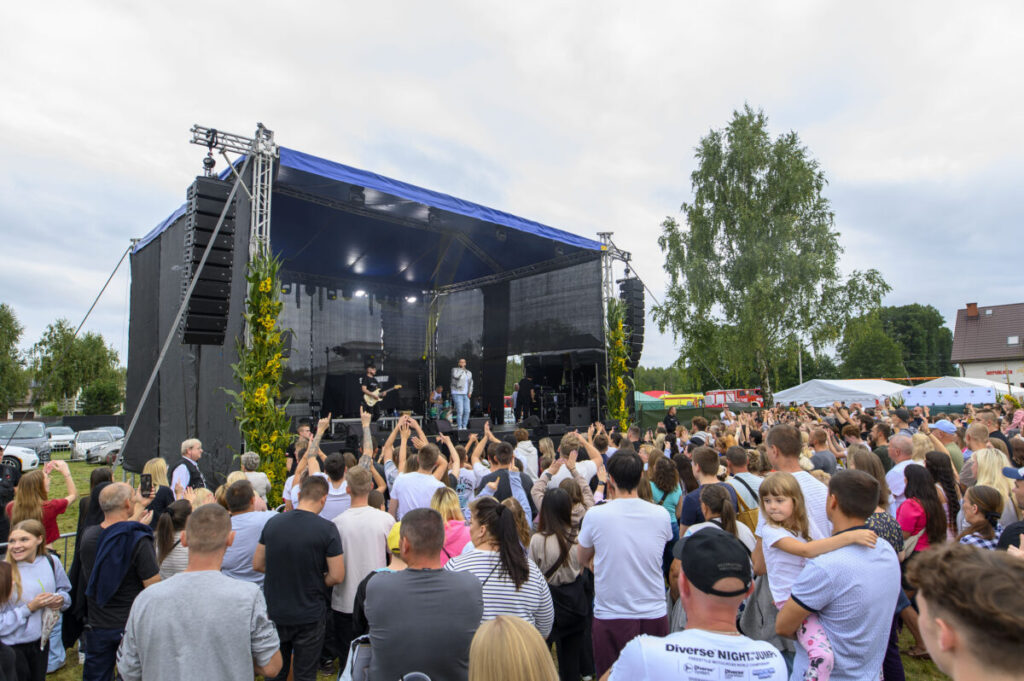 Scena na otwartym powietrzu z zespołem muzycznym, przed nią tłum ludzi. Dominują kolory niebieski, zielony i czarny. W tle drzewa i namioty.
