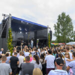 Scena na otwartym powietrzu z zespołem muzycznym, przed nią tłum ludzi. Dominują kolory niebieski, zielony i czarny. W tle drzewa i namioty.