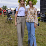 Dwie kobiety z wiankami kwiatów na głowach stoją na trawie. Ubrania w jasnych kolorach, biała bluzka i jeansy. W tle widoczne zgromadzenie ludzi i sprzęt sceniczny.