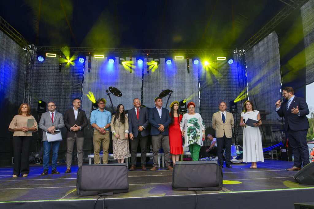 Na scenie stoi grupa ludzi w eleganckich strojach. Światła w kolorach żółtym i niebieskim oświetlają tło. Jeden z mężczyzn mówi do mikrofonu.