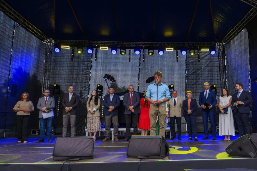 Na scenie stoi kilkanaście osób w formalnych strojach. Tło czarne, z efektami świetlnymi. Przemawia mężczyzna w błękitnej koszuli.