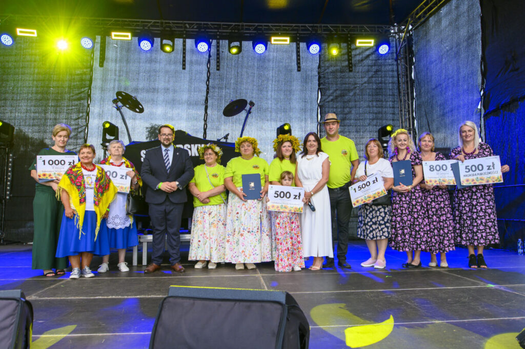 Grupa osób stoi na scenie, trzymając duże czeki. Kolory dominujące to zielony i niebieski. W tle widać ciemne oświetlenie sceny.