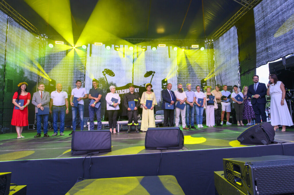 Grupa ludzi stoi na scenie oświetlonej żółtymi reflektorami. Każda osoba trzyma certyfikat. Ciemne tło wypełnia przestrzeń za nimi.