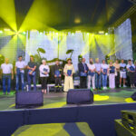 Grupa ludzi stoi na scenie oświetlonej żółtymi reflektorami. Każda osoba trzyma certyfikat. Ciemne tło wypełnia przestrzeń za nimi.