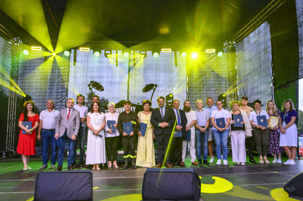 Zdjęcie przedstawia grupę ludzi na scenie w kolorowych ubraniach. W tle czarne, podświetlone kurtyny i żółte reflektory. Obecni trzymają dyplomy, co sugeruje ceremonię wręczenia nagród.