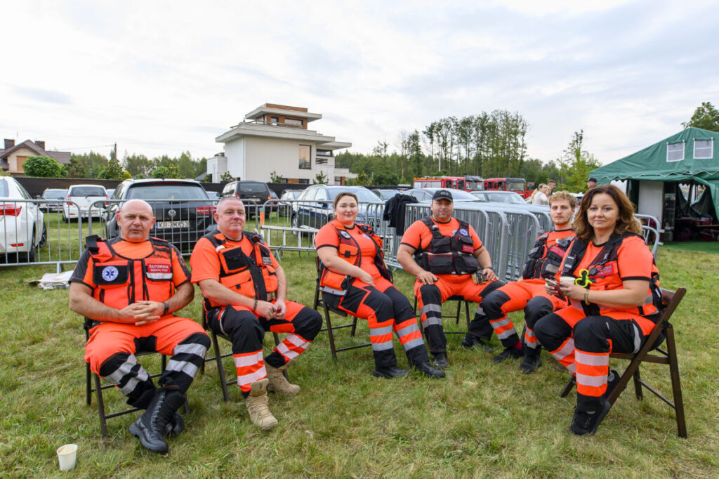 Grupa ratowników w pomarańczowych uniformach siedzi na krzesłach na trawie. W tle białe samochody i budynek. Dzień pochmurny.