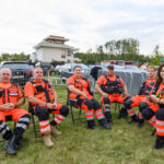 Grupa ratowników w pomarańczowych uniformach siedzi na krzesłach na trawie. W tle białe samochody i budynek. Dzień pochmurny.