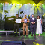 Na zdjęciu widać cztery osoby na scenie z kolorowym oświetleniem. Tło to ciemna zasłona z logo. Dwie osoby trzymają dyplomy.