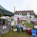 Na zdjęciu widać stoisko z obrazami na świeżym powietrzu. Kolorowe flagi zdobią zielony namiot. W tle stoi biały budynek z brązowym dachem.