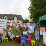 Na zdjęciu jest plenerowa wystawa sztuki z obrazami drzew. Przedstawione są kolorowe pejzaże na sztalugach, w tle jest dom i drzewa. Po lewej stronie widoczny jest biały baner z napisem Mazowsze.