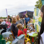 Na zdjęciu jest grupa ludzi na pikniku, z kolorowym kołem fortuny z przodu. Tło to scena z muzyką na żywo i zielone drzewa. Jest dzień, a niebo jest pochmurne.