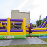 Na zdjęciu widoczny jest kolorowy, wielki, nadmuchiwany plac zabaw z fioletowymi, żółtymi i zielonymi elementami. Dzieci bawią się na torze przeszkód w obszarze na trawie. W tle znajduje się budynek oraz drzewa pod