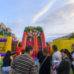 Na zdjęciu widoczna jest duża, kolorowa zjeżdżalnia dmuchana z motywem zwierząt. W tle widać budynki i błękitne niebo. Wokół stoi grupa ludzi obserwujących zabawę dzieci.