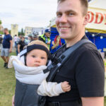 Mężczyzna trzymający dziecko w nosidełku podczas plenerowego wydarzenia. Jasne, pastelowe kolory i tłum w tle. Pogodny nastrój.