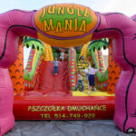 Kolorowa dmuchana zjeżdżalnia w kształcie różowych słoni, z napisem Jungle Mania. Dzieci bawią się na czerwono-żółtym tle. W tle widoczne są zielone elementy przypominające dżunglę.