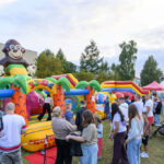 Na zdjęciu widać kolorowe dmuchane zamki w kształcie jungli z małpą na szczycie. Wokół stoją ludzie w kolejce. W tle są zielone drzewa i kolorowe namioty.