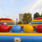 Dzieci bawią się na kolorowym dmuchanym zamku z dużymi kulami w czerwieni, żółci i błękicie. Tło stanowią drzewa i budynek. Nad nimi jasne, zachmurzone niebo.